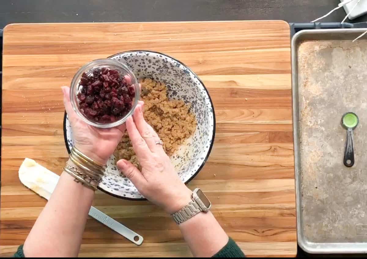 A person’s hands hold a small bowl of dried cranberries over a larger mixing bowl containing cookie dough; a baking sheet and measuring spoon are on the wooden countertop.