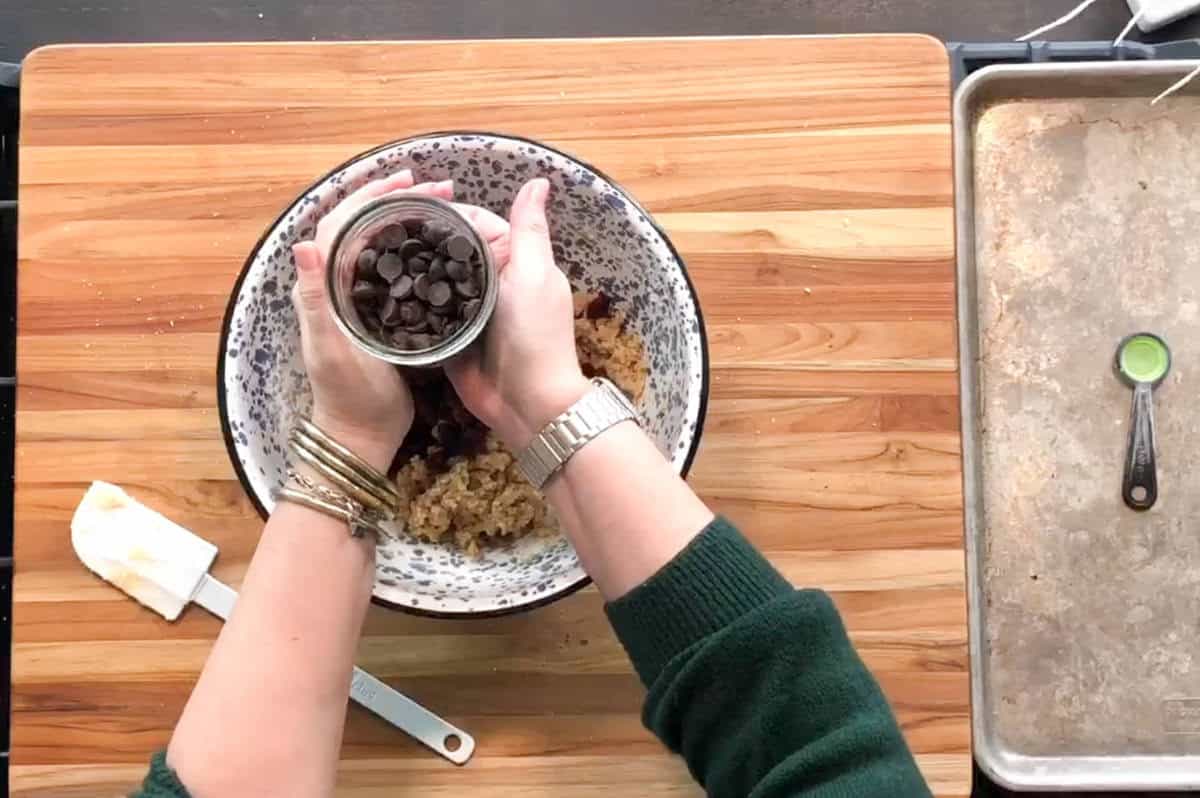 A person wearing a green sweater pours chocolate chips from a small glass jar into a large mixing bowl with cookie dough, on a wooden countertop next to a spatula, measuring spoon, and baking sheet.
