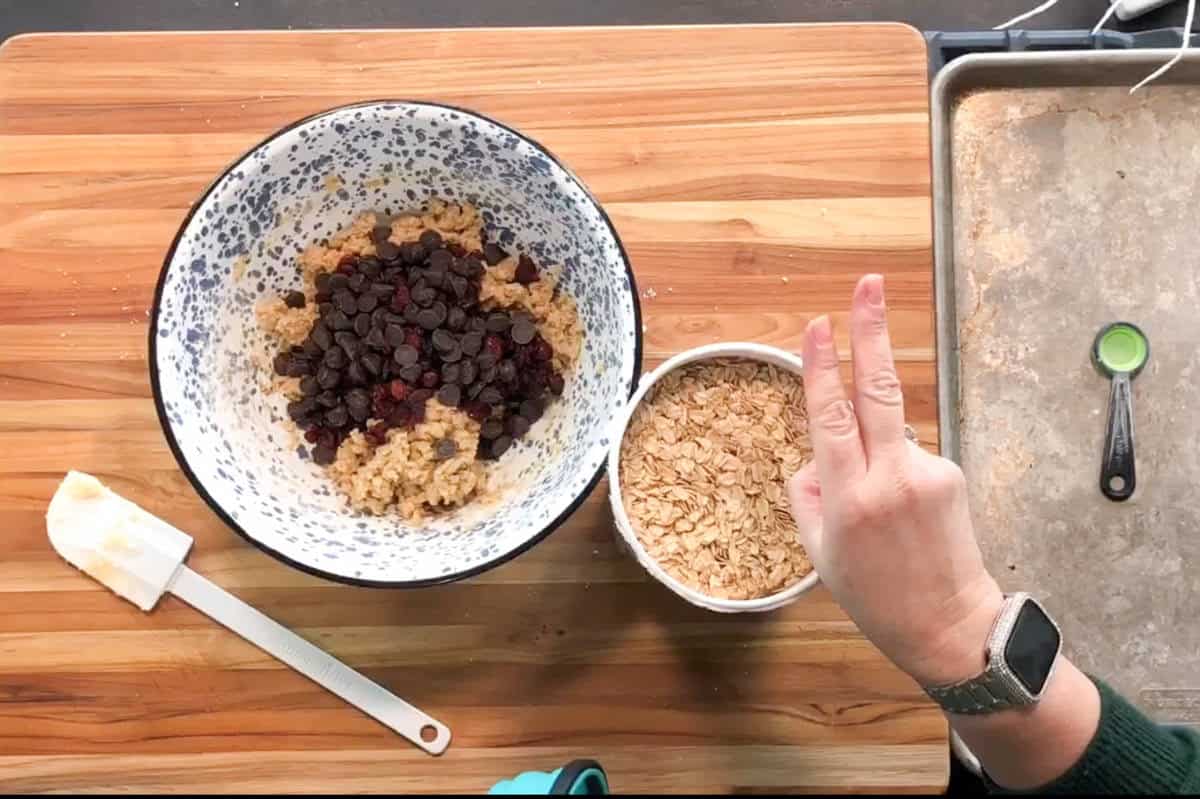 A person holds up two fingers over a wooden counter with a mixing bowl of cookie dough, dried fruit, and chocolate chips, a small bowl of oats, a spatula, a measuring spoon, and a baking sheet.