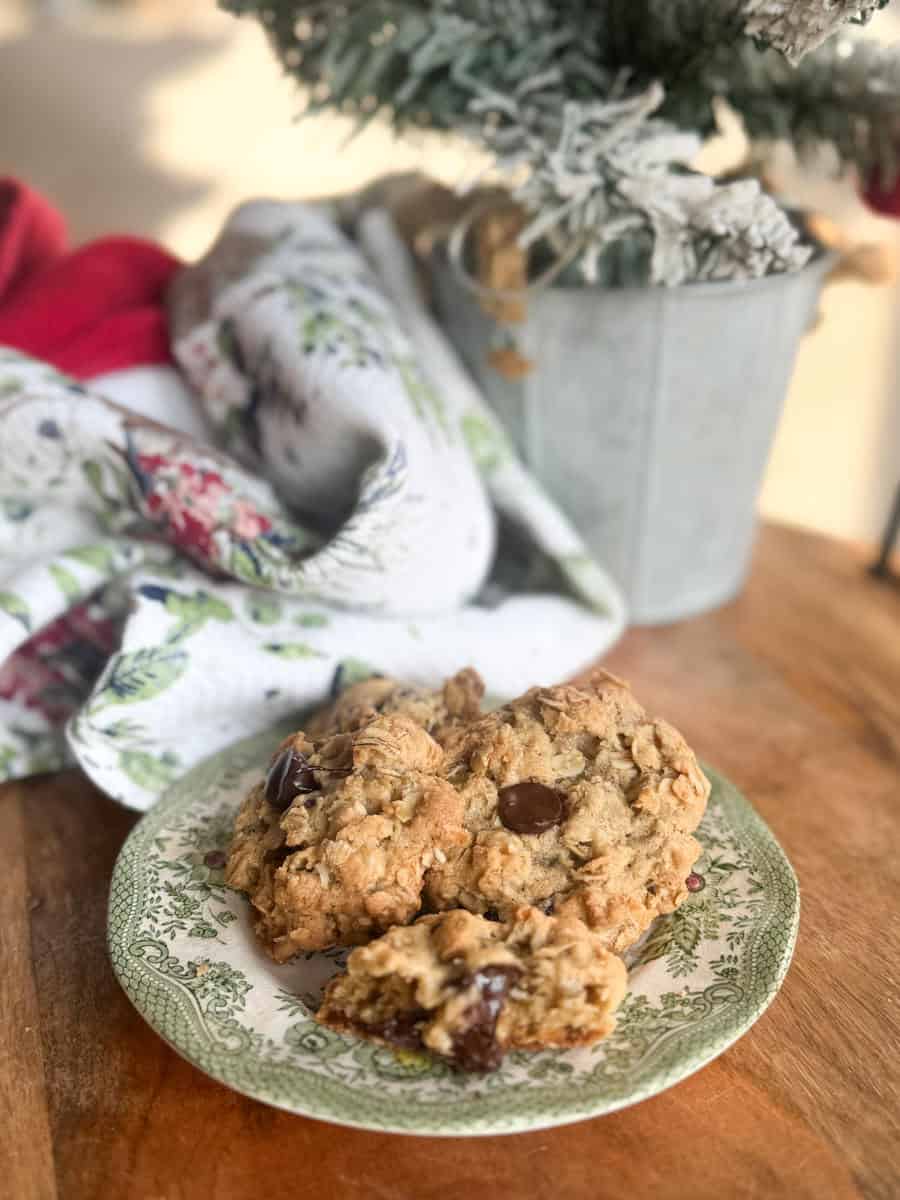 A green and white patterned plate holds a large, chunky cookie with visible chocolate chips. The plate is on a wooden surface near a floral cloth and a small potted plant with snow-dusted leaves.