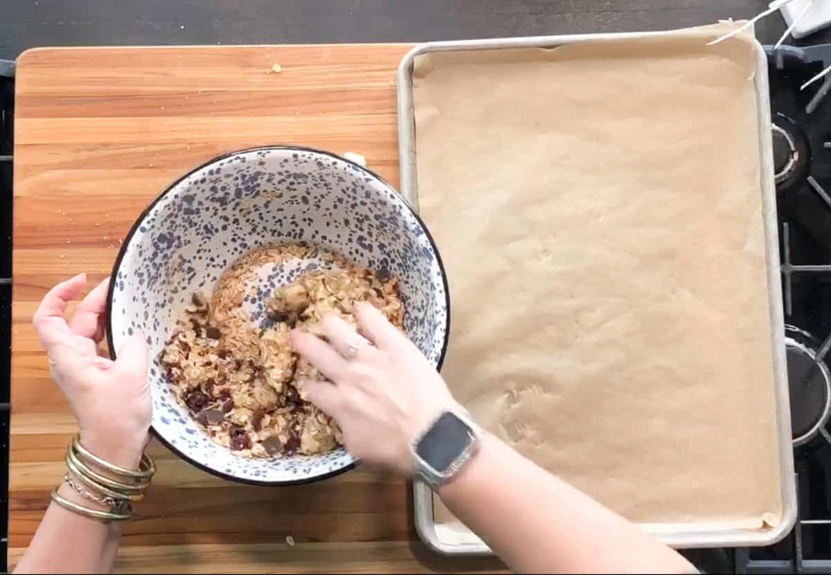 A person mixes ingredients by hand in a large bowl on a wooden surface next to a baking sheet lined with parchment paper. The person is wearing several bracelets and a smartwatch.