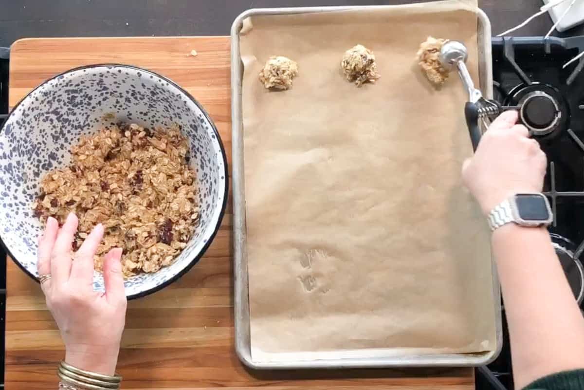 A person uses a scoop to place balls of oatmeal cookie dough onto a parchment-lined baking sheet; a large bowl of dough is on the left on a wooden surface.