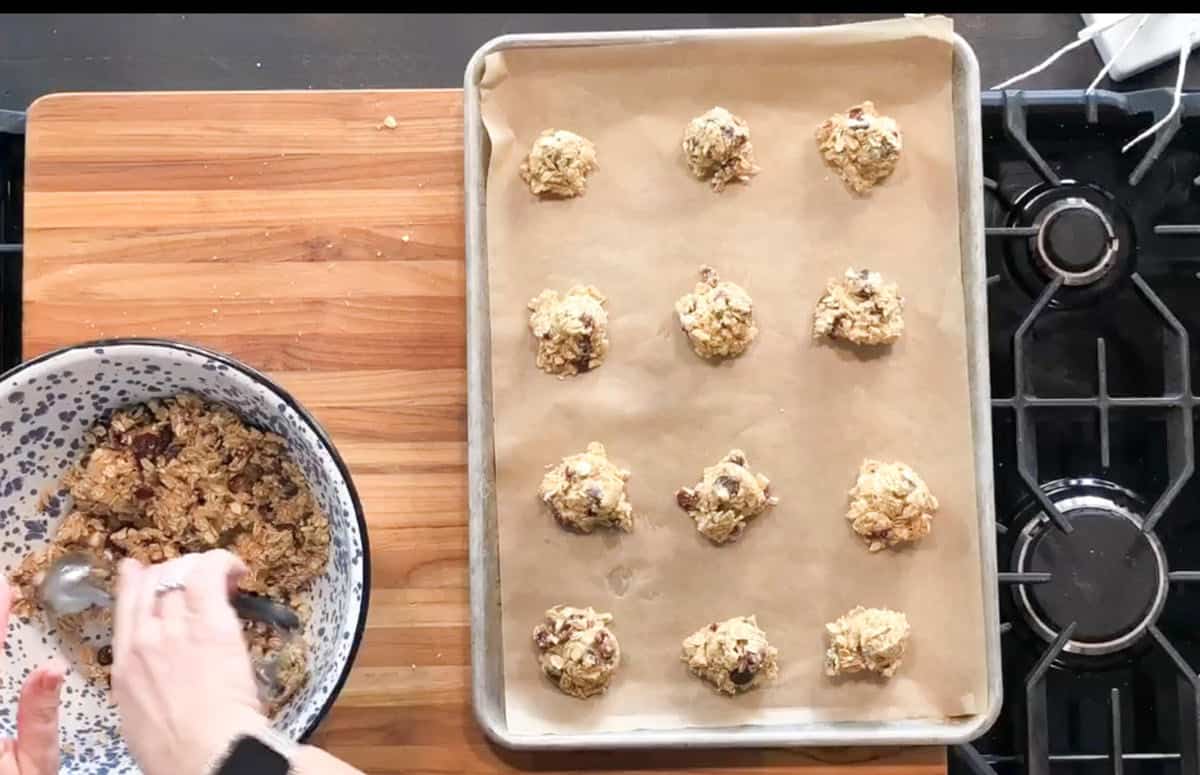 A person scoops cookie dough from a large bowl onto a baking sheet lined with parchment paper. The baking sheet holds 12 evenly spaced dough portions, and both items are on a wooden countertop next to a stove.
