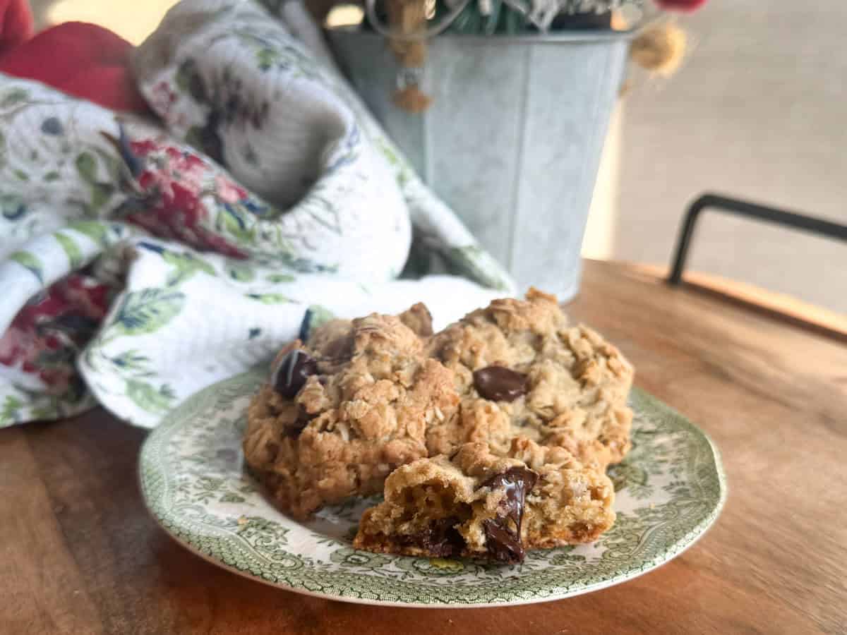 A green and white patterned plate holds two chocolate chip oatmeal cookies, one partially broken to show melted chocolate. The plate is on a wooden table near a floral towel and a metal planter in the background.