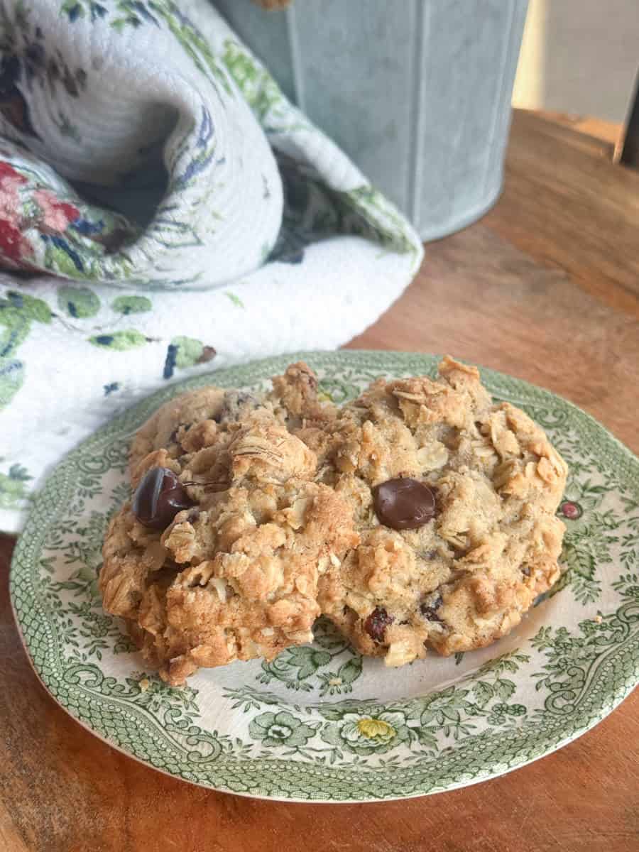 Two oatmeal cookies with chocolate chips and raisins are placed on a vintage green-patterned plate. A white towel with a green and red floral design is partially visible in the background on a wooden surface.