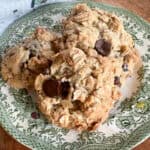 Three large oatmeal chocolate chip cookies are stacked on a decorative, green floral-patterned plate. The cookies have a chunky texture with visible oats and chocolate chips. The plate is placed on a wooden surface.
