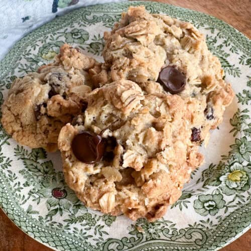 Three large oatmeal chocolate chip cookies are stacked on a decorative, green floral-patterned plate. The cookies have a chunky texture with visible oats and chocolate chips. The plate is placed on a wooden surface.