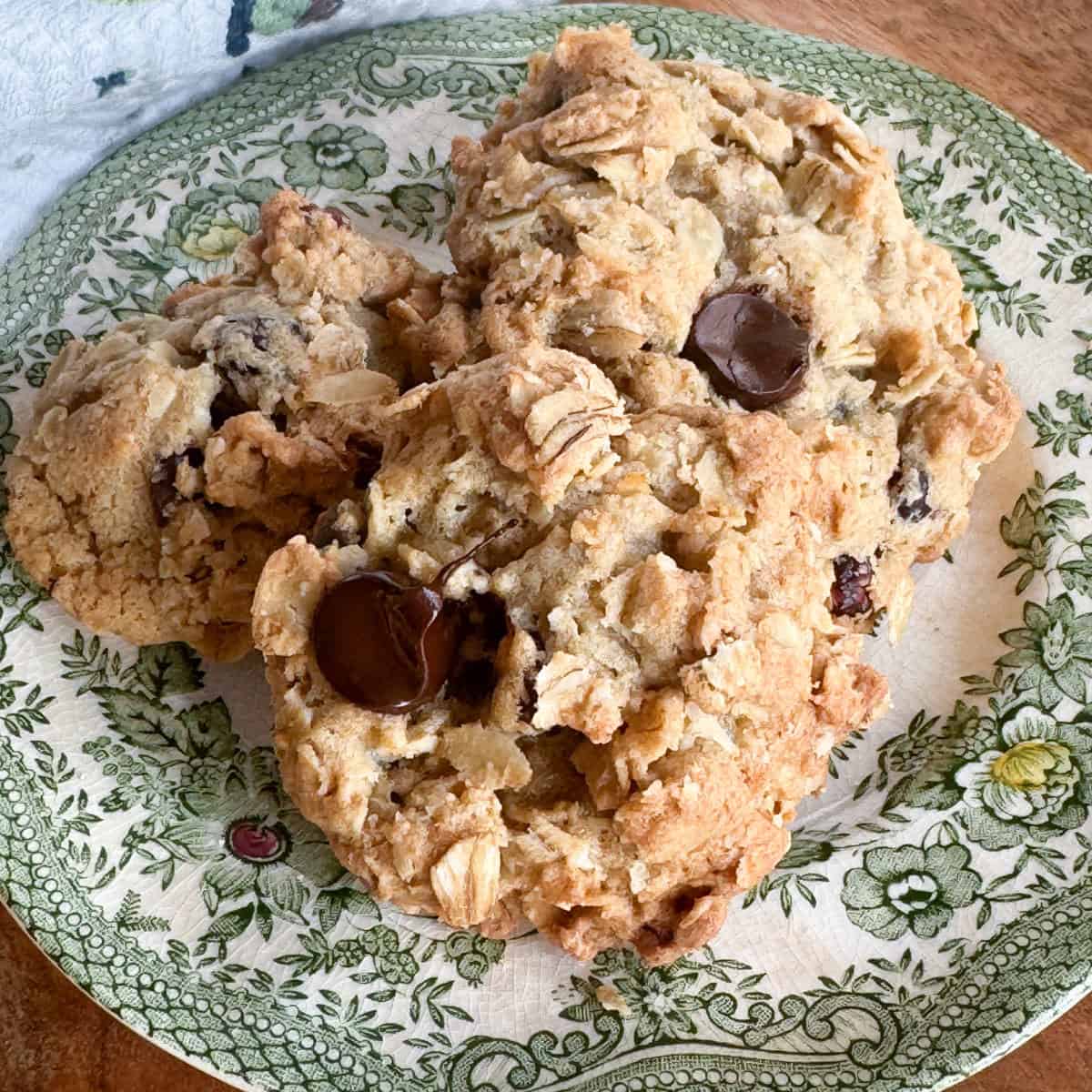 Three large oatmeal chocolate chip cookies are stacked on a decorative, green floral-patterned plate. The cookies have a chunky texture with visible oats and chocolate chips. The plate is placed on a wooden surface.