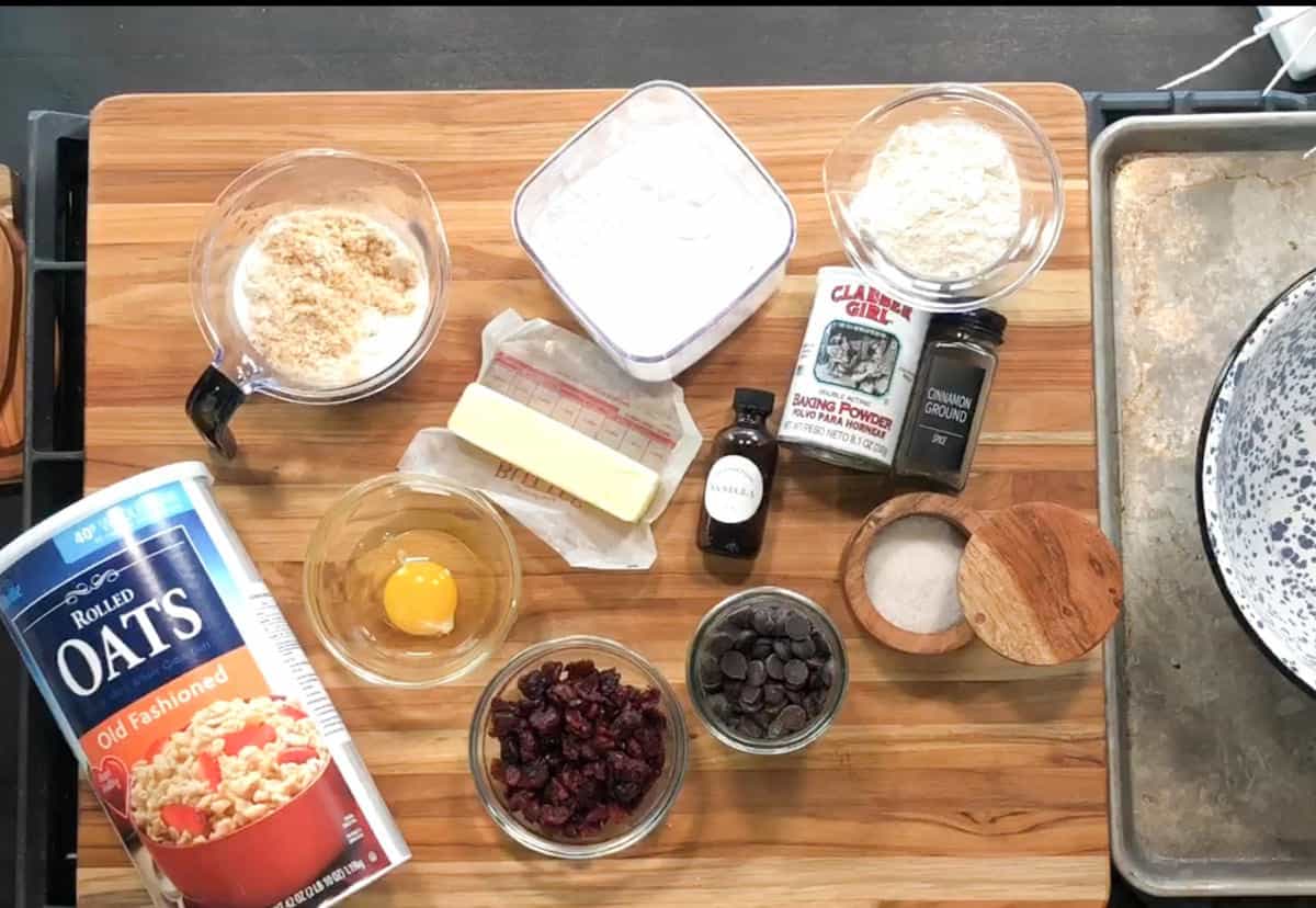A variety of baking ingredients arranged on a wooden surface, including oats, an egg, butter, flour, brown sugar, chocolate chips, dried cranberries, vanilla extract, baking powder, cinnamon, and salt.