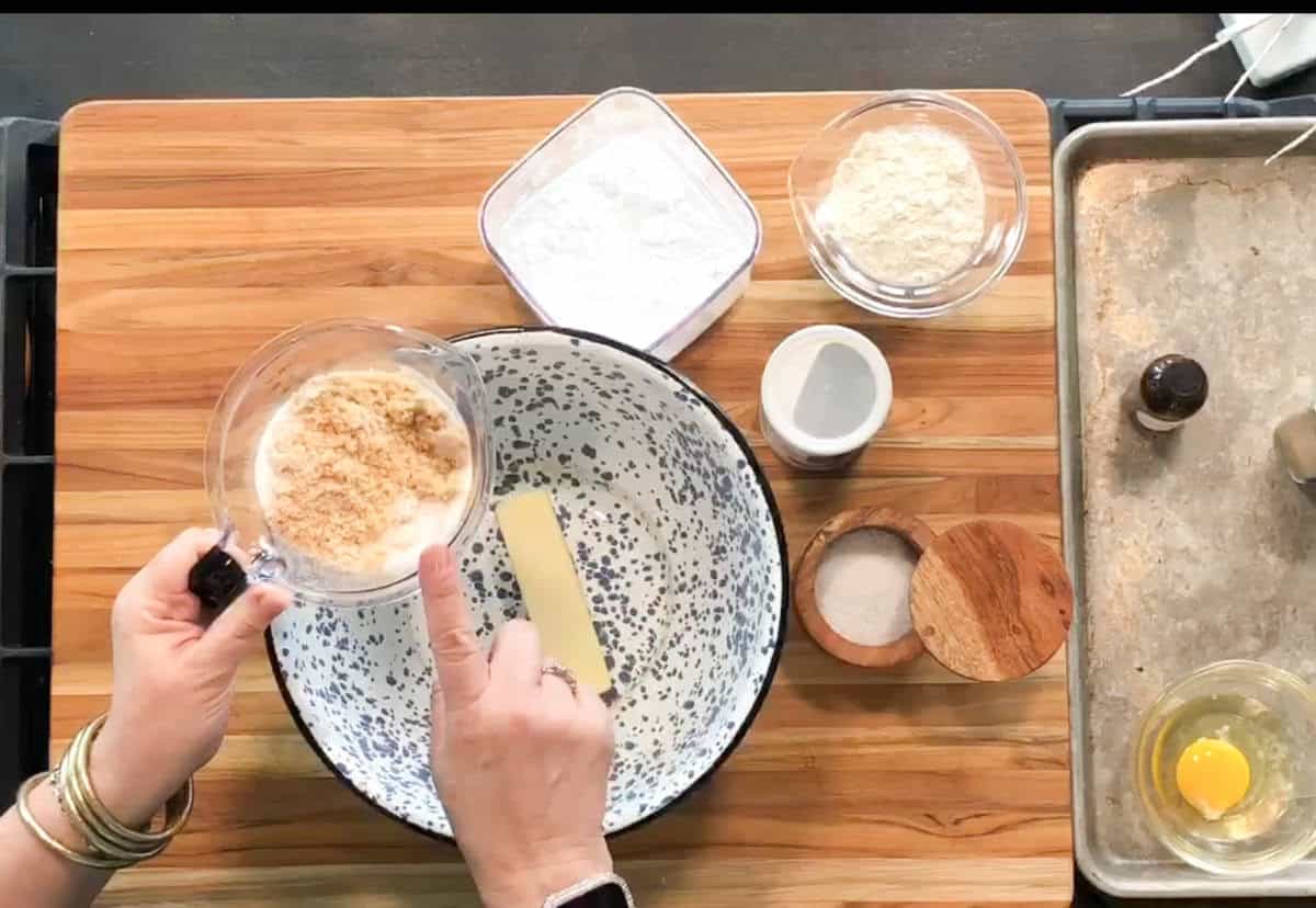 A person holds a measuring cup of brown sugar above a mixing bowl with a stick of butter inside. Other ingredients, including flour, powdered sugar, salt, an egg, and vanilla, are arranged on a wooden surface.
