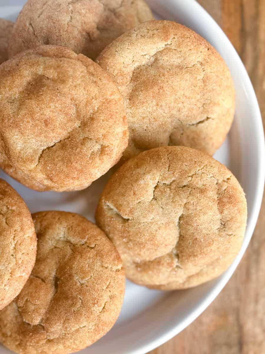 Several round, golden-brown cookies with a cracked surface are arranged closely together on a white plate. The texture appears slightly crisp on the outside. The plate is set on a wooden surface.