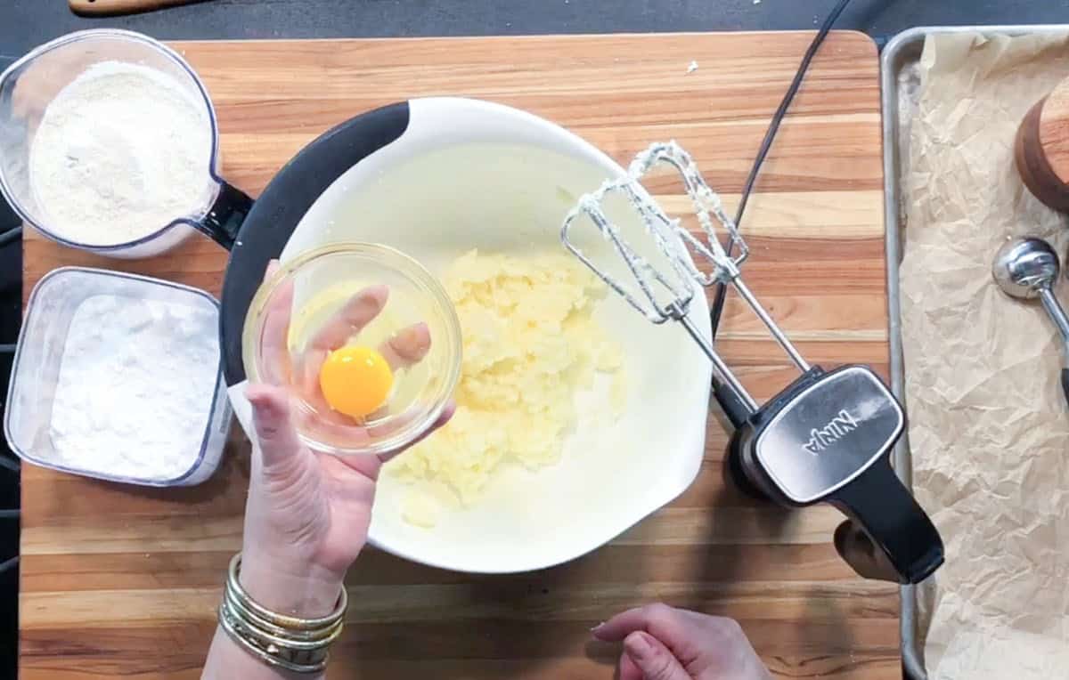 A person holds a small glass bowl with a cracked egg above a mixing bowl containing a creamed mixture. An electric hand mixer, a container of flour, and a container of powdered sugar are nearby on a wooden surface.