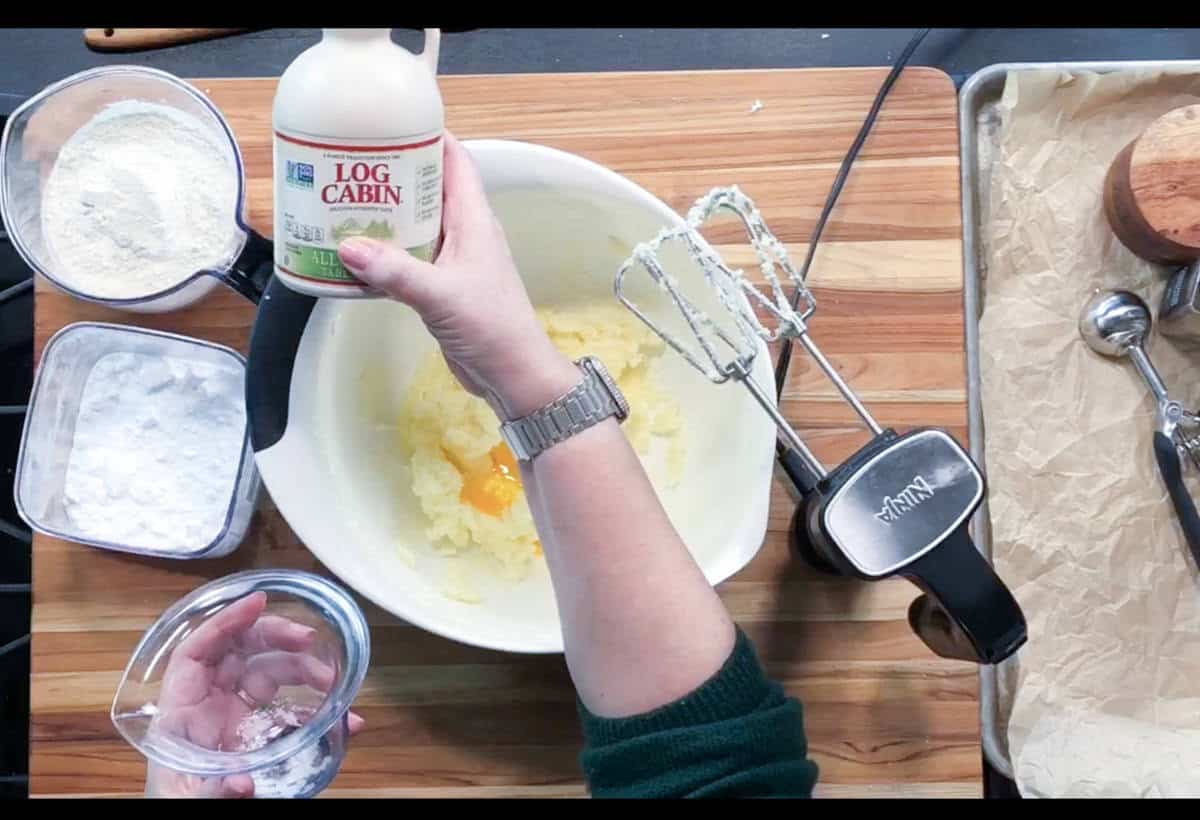 A person holds a bottle of Log Cabin syrup and a measuring cup over a mixing bowl with eggs and batter. An electric mixer, flour, sugar, and baking tools are on a wooden surface beside parchment-lined pan.