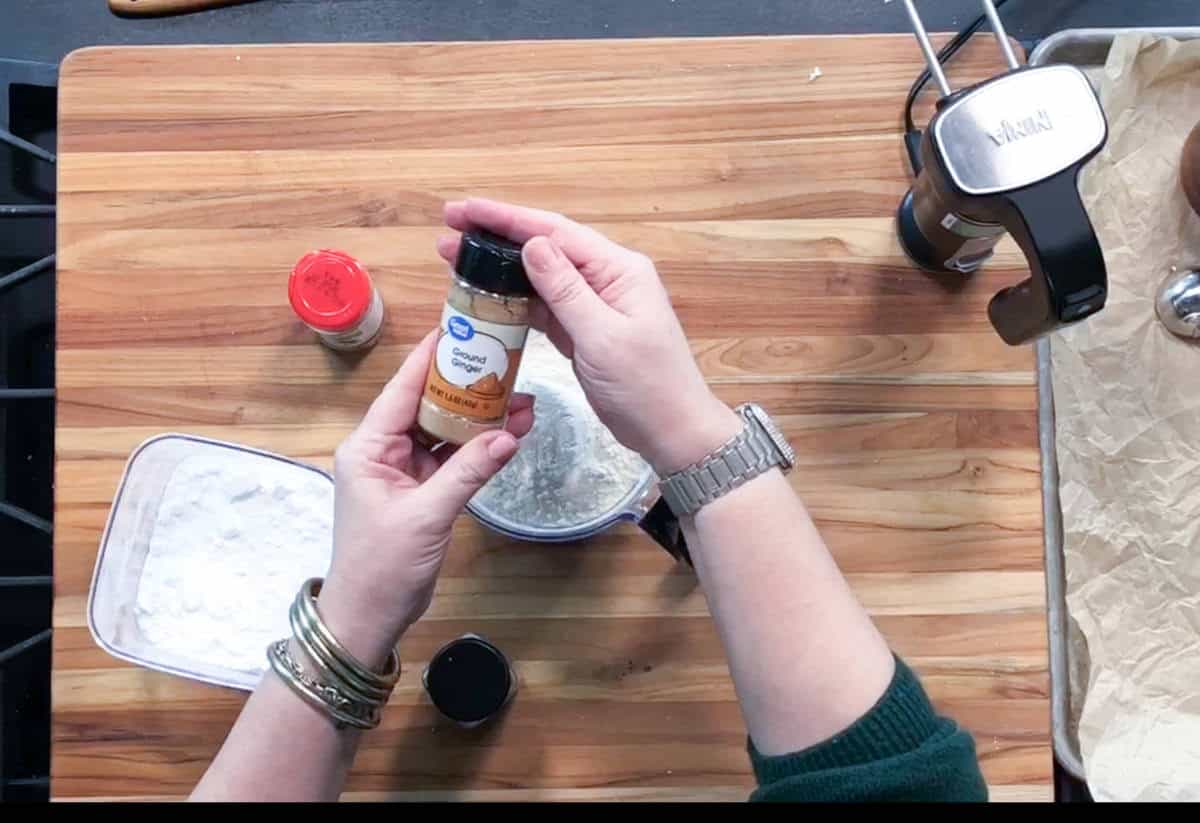 A person holds a container of ground ginger above a mixing bowl filled with a white powder, likely flour, on a wooden countertop. Other ingredients and a stand mixer are also visible.