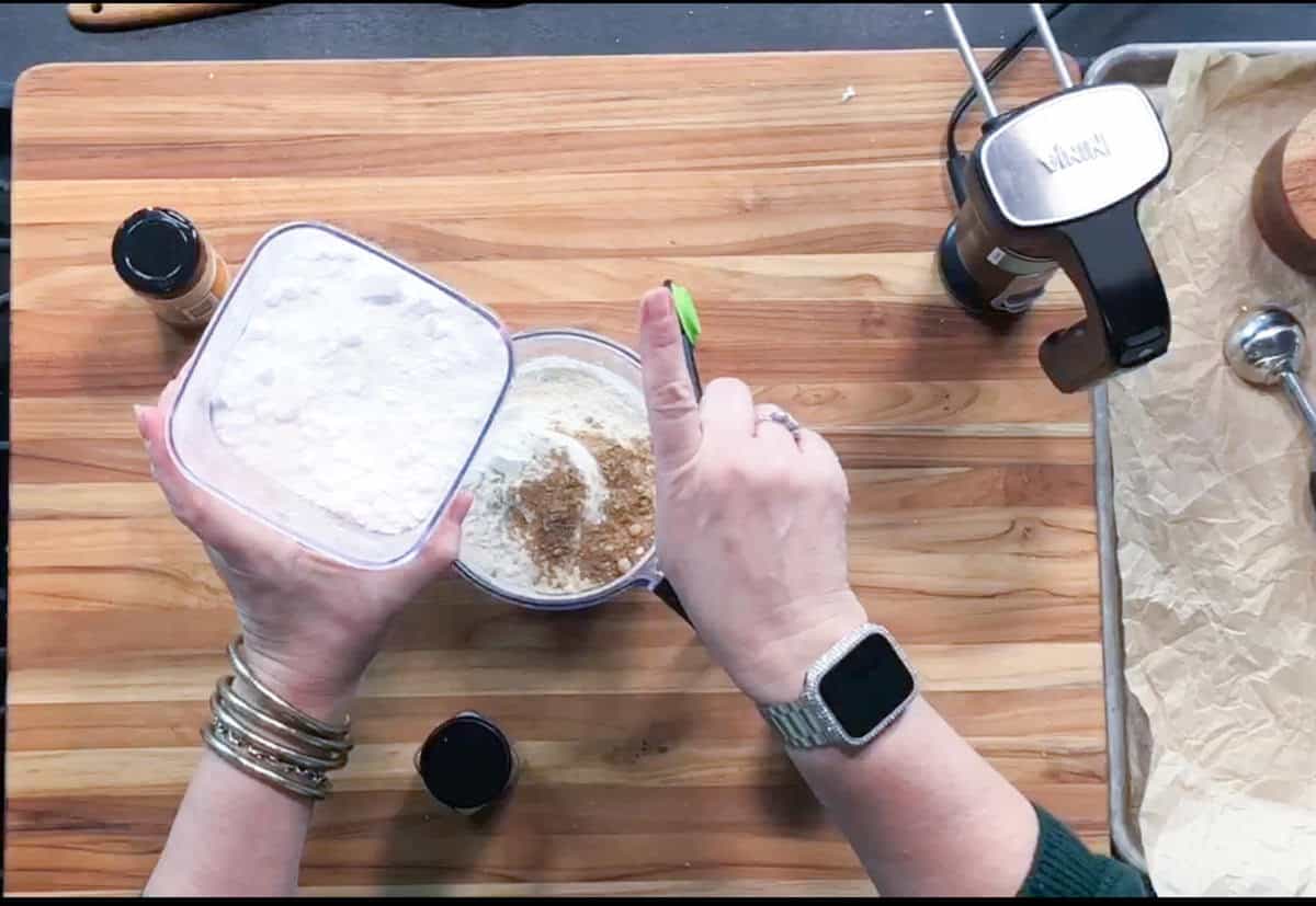 A person wearing a smartwatch pours a container of flour or powdered sugar into a bowl with spices on a wooden cutting board, alongside spice jars, a stand mixer, and measuring spoons.
