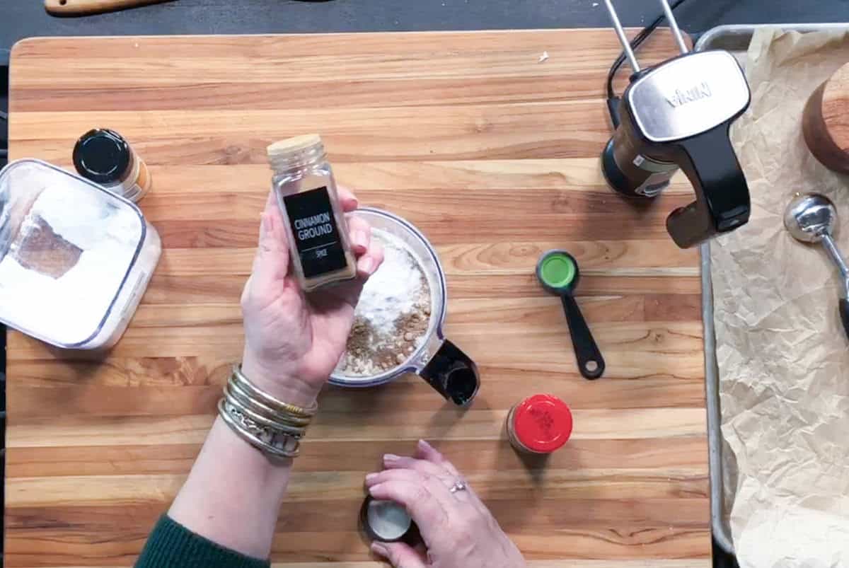 A person holding a bottle of ground cinnamon above a mixing bowl on a wooden countertop, surrounded by measuring spoons, containers, and a stand mixer.