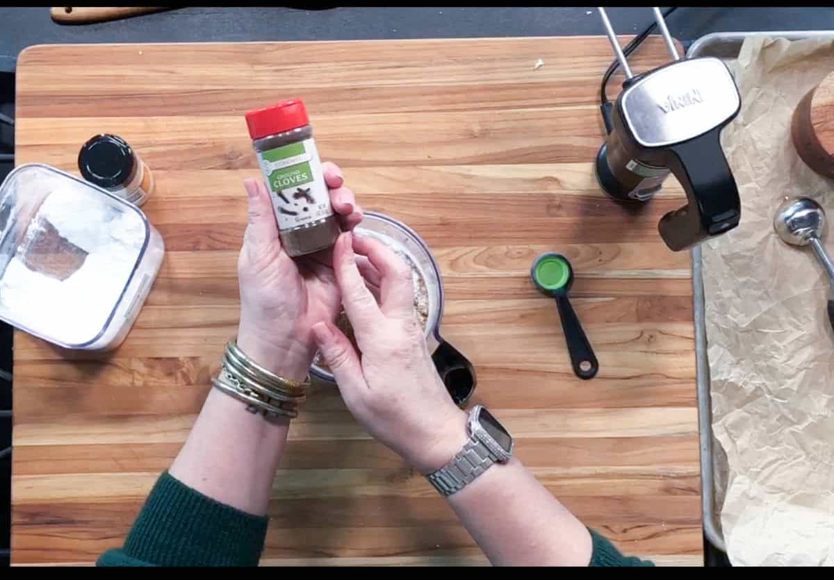 A person holding a container of ground cloves over a wooden cutting board, with kitchen tools, a teaspoon, a food processor, and ingredient containers visible on the counter.