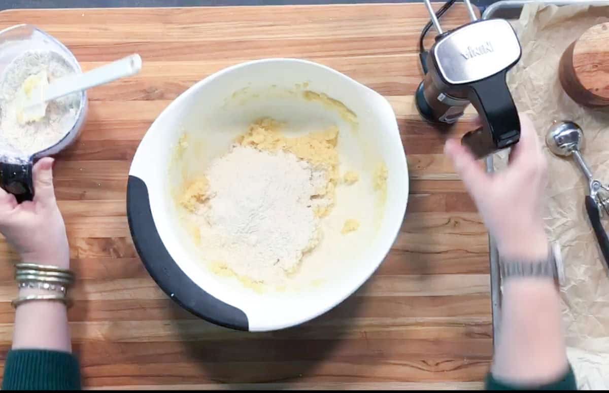 A person adds dry ingredients from a measuring cup into a bowl of yellow batter on a wooden countertop, with a stand mixer and various utensils nearby.