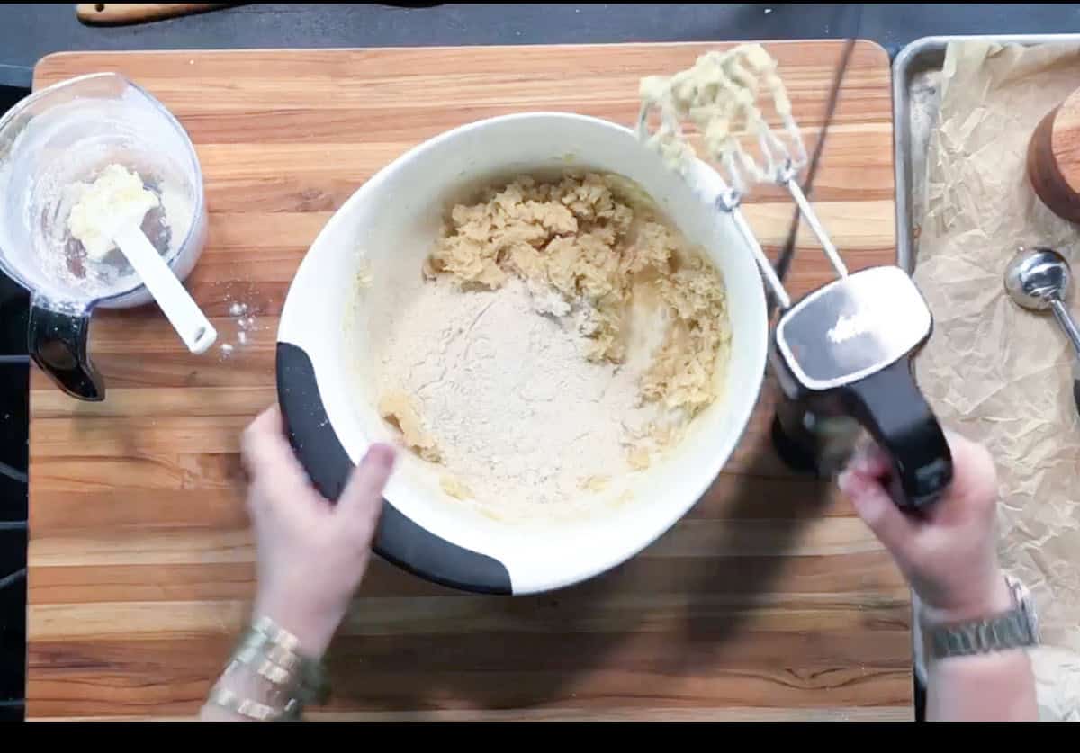 A person uses a hand mixer to blend flour into dough in a large white mixing bowl. A spatula in a smaller bowl with dough residue is nearby, and a parchment-lined baking sheet is on the right.