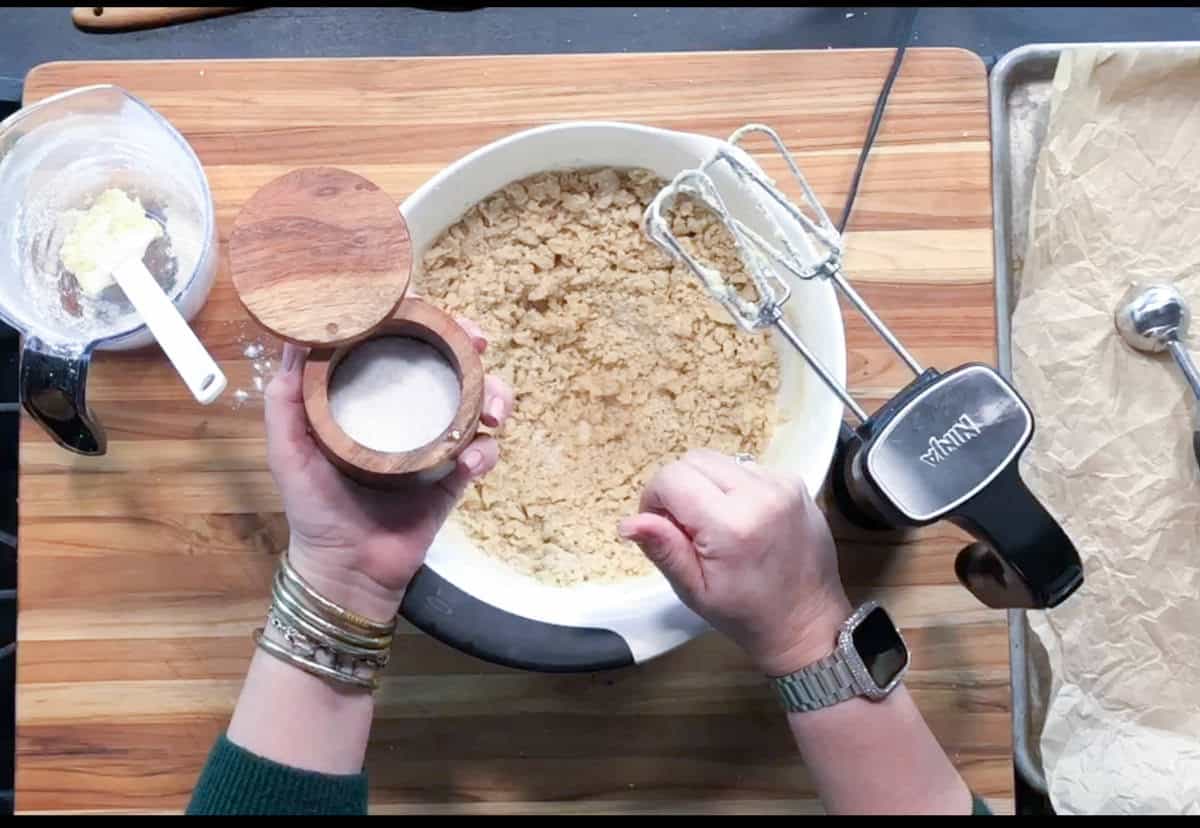 Person holding a wooden salt cellar over a mixing bowl with cookie dough, next to a stand mixer, a bowl with butter, and a baking tray lined with parchment paper on a wooden surface.