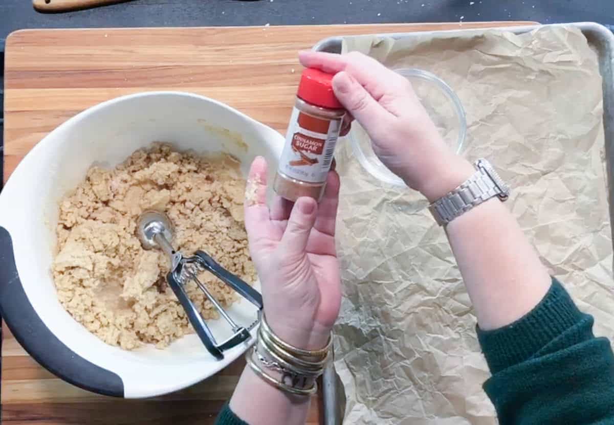 A person holds a container of ground cinnamon above a tray lined with parchment paper. A mixing bowl with cookie dough and a cookie scoop are nearby on a wooden surface.