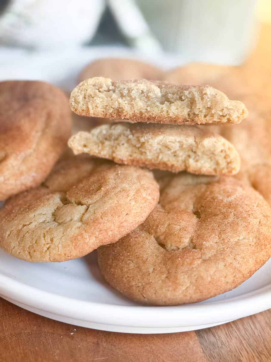 A white plate with several golden brown cookies. One cookie is broken in half and stacked on top, showing its soft, crumbly interior. The plate sits on a wooden surface.