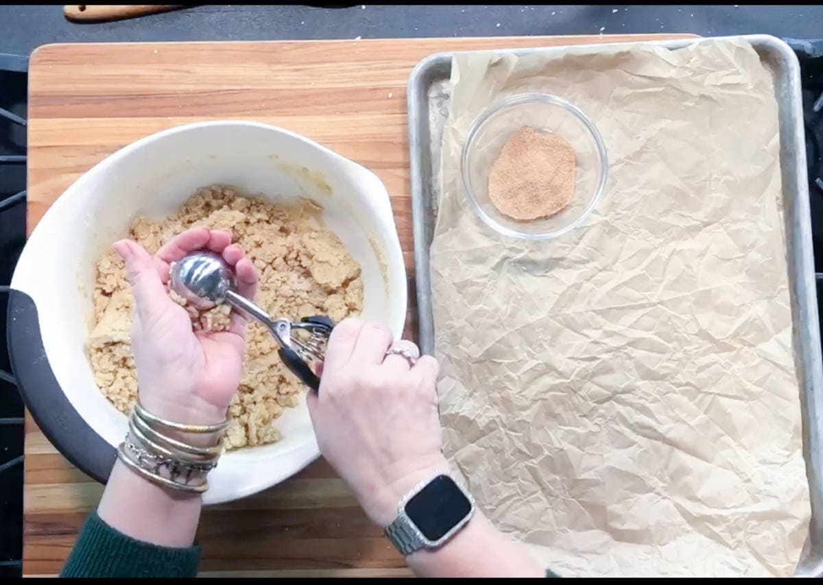 A person holding a cookie scoop over a bowl of dough, preparing to form cookies. A tray lined with parchment paper and a small bowl of cinnamon sugar sit on a wooden surface nearby.