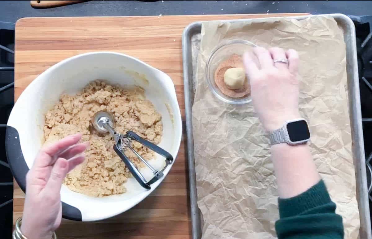 A person rolls cookie dough in cinnamon sugar beside a mixing bowl of dough and a lined baking sheet. A cookie scoop rests in the bowl, and the person wears a watch on their left wrist.