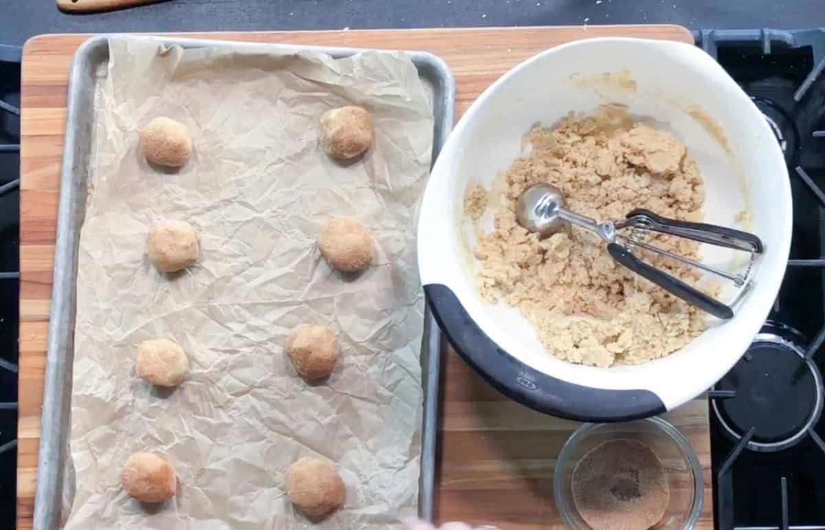 A baking tray with eight cookie dough balls on parchment paper sits beside a white mixing bowl containing more dough and a cookie scoop, on a wooden surface near a stove.