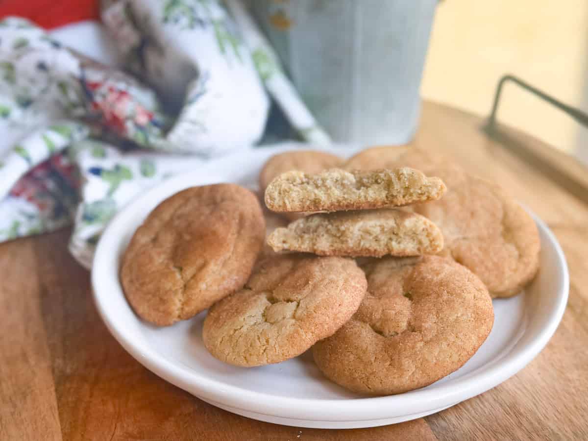 A white plate on a wooden surface holds five snickerdoodle cookies, with one cookie broken in half to show the inside. A floral napkin and a metal container are in the background.