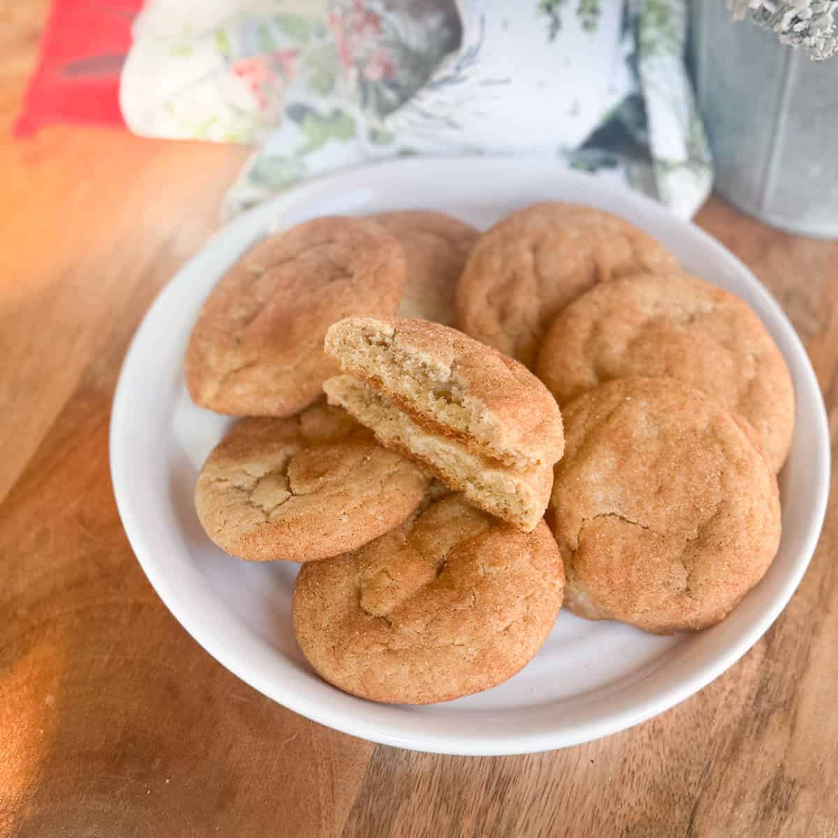 A white plate filled with several cinnamon sugar cookies sits on a wooden surface. One cookie is broken in half, revealing a soft, textured interior. A patterned cloth is partially visible in the background.