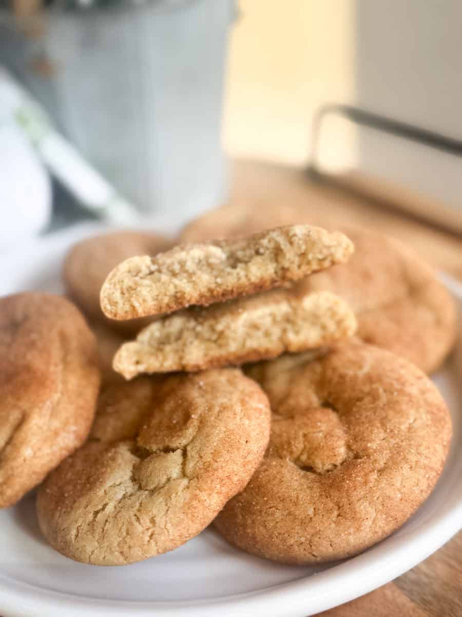 A plate of round snickerdoodle cookies, with one cookie broken in half and placed on top to show the soft, textured interior. The background is slightly blurred.