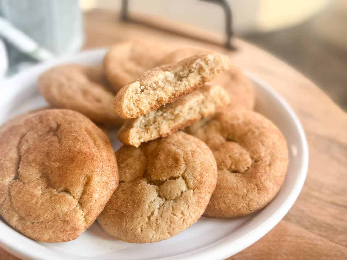 A white plate holds several round, golden-brown cookies. One cookie is broken in half, showing a soft, crumbly interior. The plate is on a wooden surface.