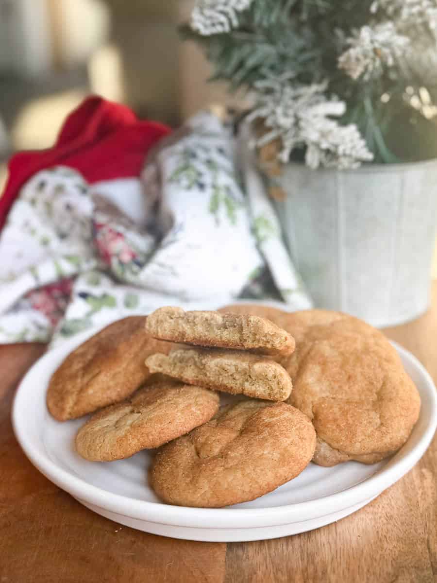 A white plate holds four snickerdoodle cookies, one broken in half to show the inside. A festive cloth and a potted plant with frosted branches are in the blurred background.