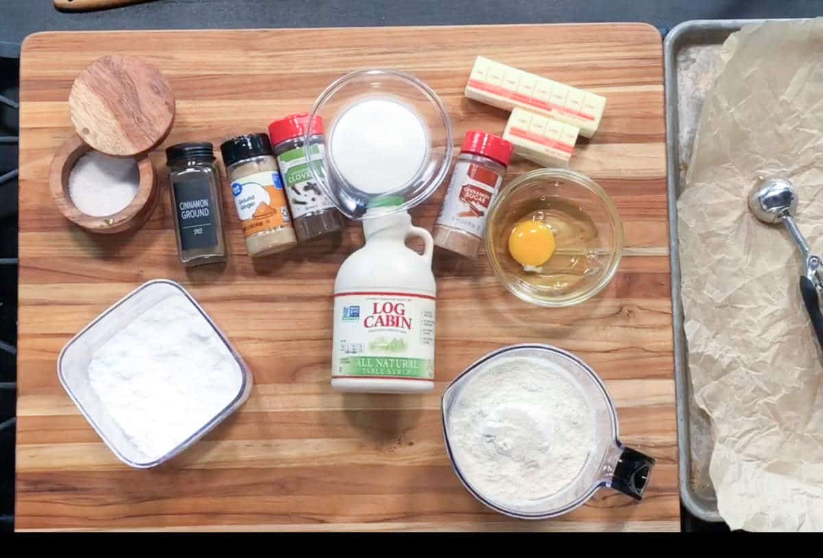 A wooden cutting board displays baking ingredients: ground cinnamon, other spices, sugar, vanilla extract, sticks of butter, an egg in a glass bowl, flour, powdered sugar, maple syrup, and a metal cookie scoop on a tray.