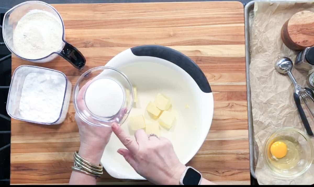 A person holds a small bowl of granulated sugar above a mixing bowl with cubed butter. Other ingredients like flour, powdered sugar, and an egg are arranged on a wooden counter.
