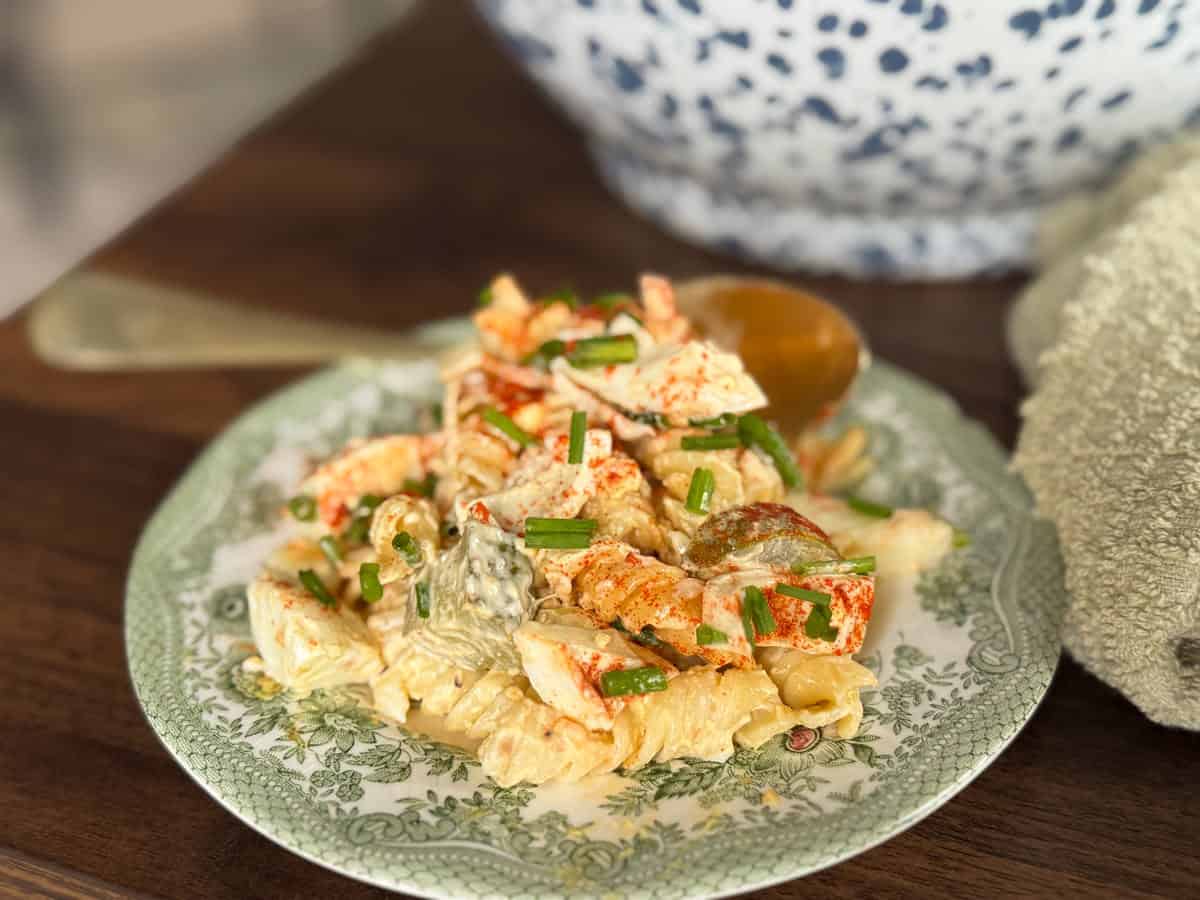 A green floral plate holds a serving of creamy pasta with pieces of lobster and chopped chives. A gold spoon rests beside the food, and a patterned bowl is partially visible in the background.