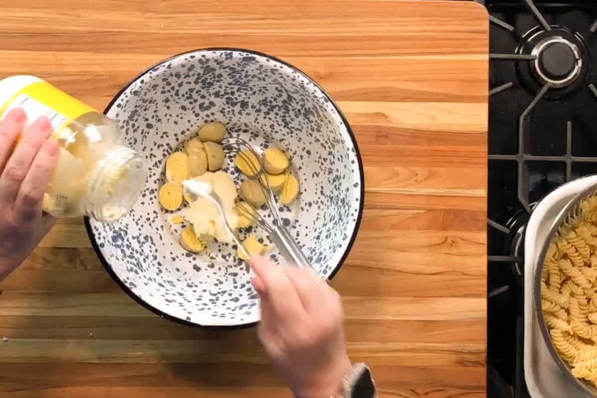 A person adds a spoonful of mayonnaise from a jar to sliced yellow potatoes in a speckled mixing bowl on a wooden countertop. Cooked rotini pasta sits in a tray near a stovetop on the right.