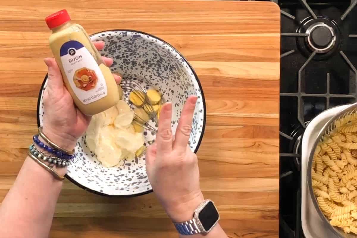 A person holds a bottle of Dijon mustard over a bowl with mayonnaise and garlic while showing a peace sign with their other hand; cooked pasta sits in a pot on a nearby stove.