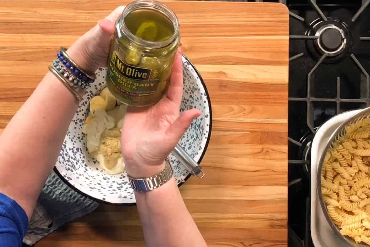 A person holds an open jar of Mt. Olive kosher baby dill pickles over a mixing bowl with sliced pickles and pasta, next to a pot of cooked rotini on a wooden countertop and a stove.