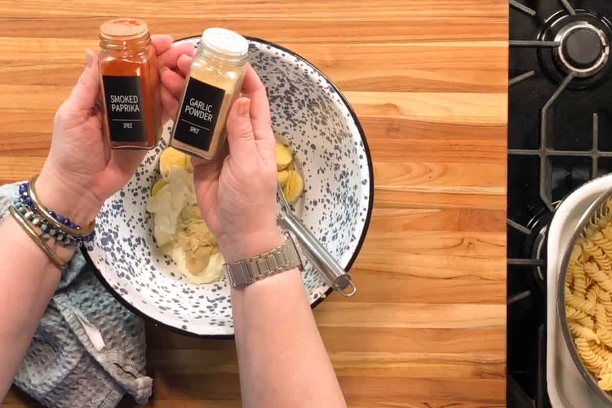 A person holding jars of smoked paprika and garlic powder above a mixing bowl with ingredients, next to a stove and a tray of cooked spiral pasta on a wooden countertop.