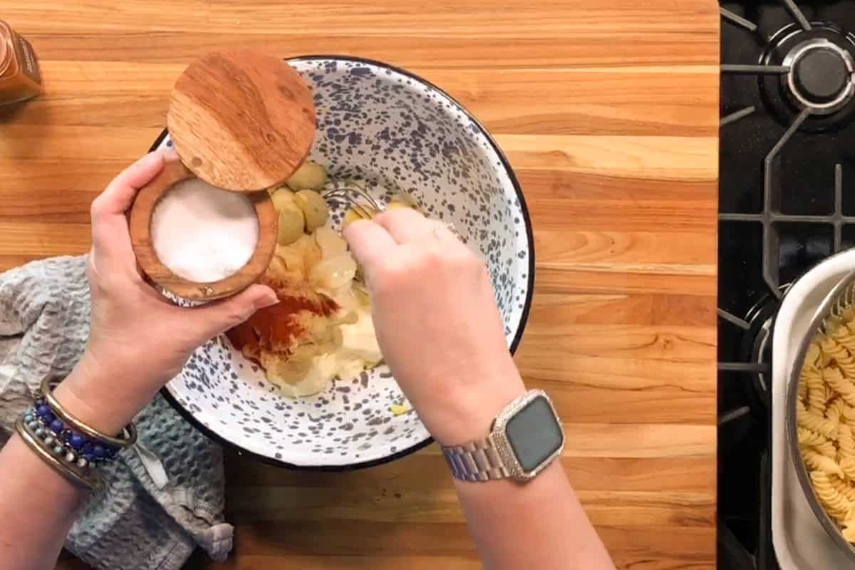 A person adds salt to a bowl containing pasta salad ingredients such as mayonnaise, spices, and pickles on a wooden countertop next to a stove and a pot of cooked rotini pasta.