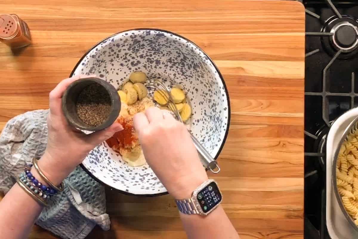 A person adds black pepper from a metal container to a large bowl containing sliced yellow vegetables, powdered spices, and a whisk. A grey towel is on the left, and cooked pasta is on the stove at the right side of the image.