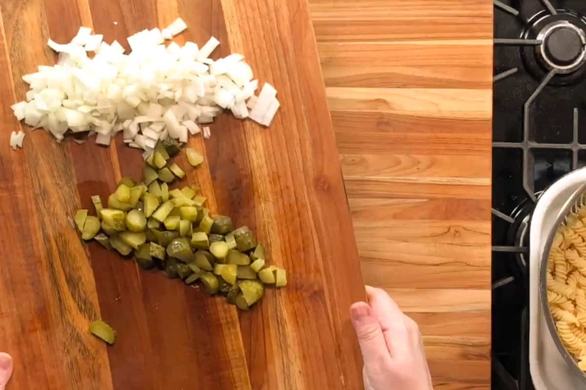A person holds a wooden cutting board with chopped onions and diced pickles above a pot of fusilli pasta on a stove. The board and stove rest on a wood countertop.