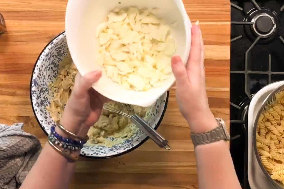 A person holds a white bowl filled with sliced onions above a mixing bowl on a wooden countertop. A metal spoon is in the mixing bowl, and cooked pasta is visible on the right by a gas stove.