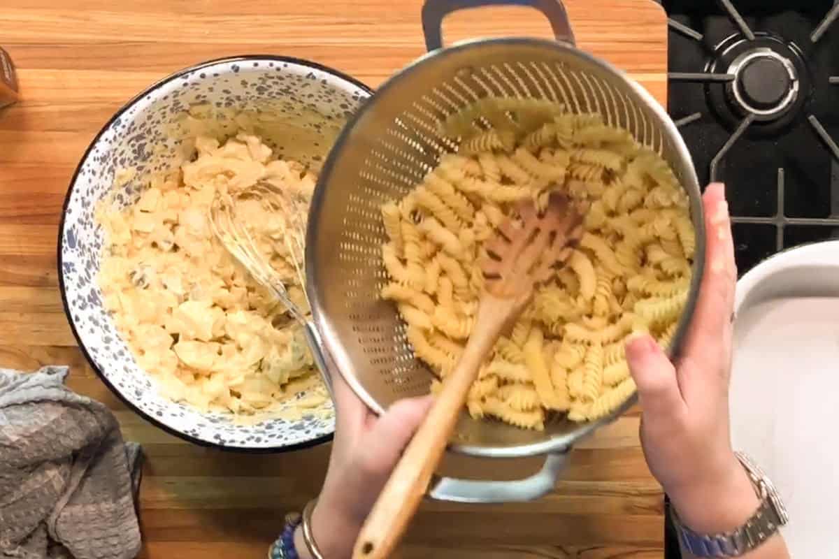 A person holds a colander of cooked rotini pasta over a wooden counter, ready to add it to a bowl of creamy sauce. A whisk, stove, and kitchen towel are also visible.