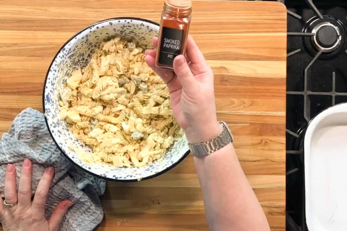 A person holds a small jar labeled "smoked paprika" above a large bowl of mixed, chopped ingredients on a wooden countertop. One hand holds a towel on the side of the bowl. Part of a stove is visible.
