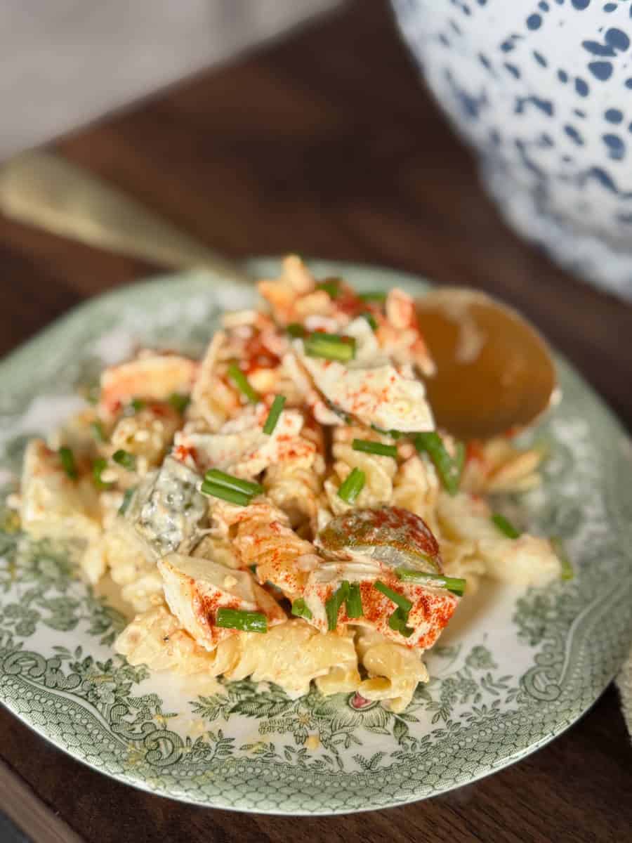 A close-up of pasta salad with chunks of lobster, chopped green onions, and paprika, served on a green and white patterned plate with a gold spoon in the background.