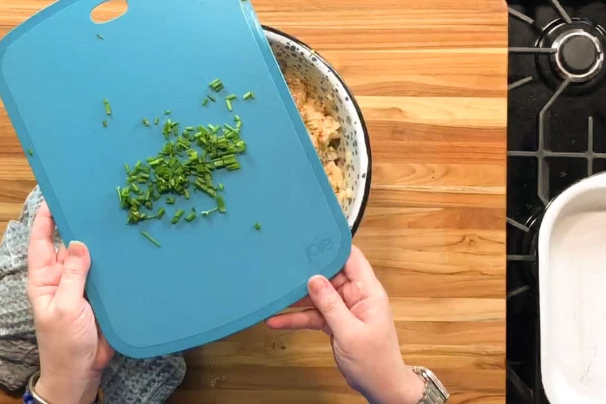 A person holds a blue cutting board with chopped green herbs above a bowl of food on a wooden countertop, next to a stove and an empty white baking dish.