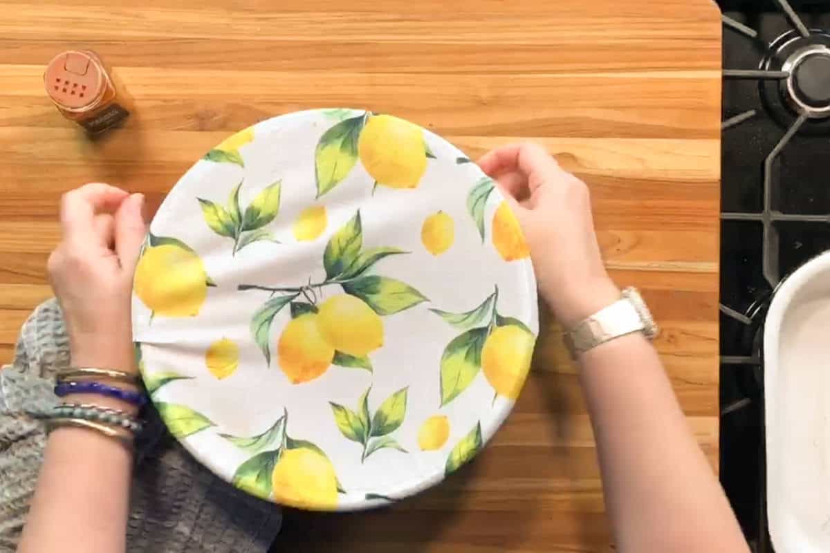 A person places a round cloth with a lemon pattern over a bowl on a wooden countertop. A jar of seasoning and part of a stovetop are visible nearby.
